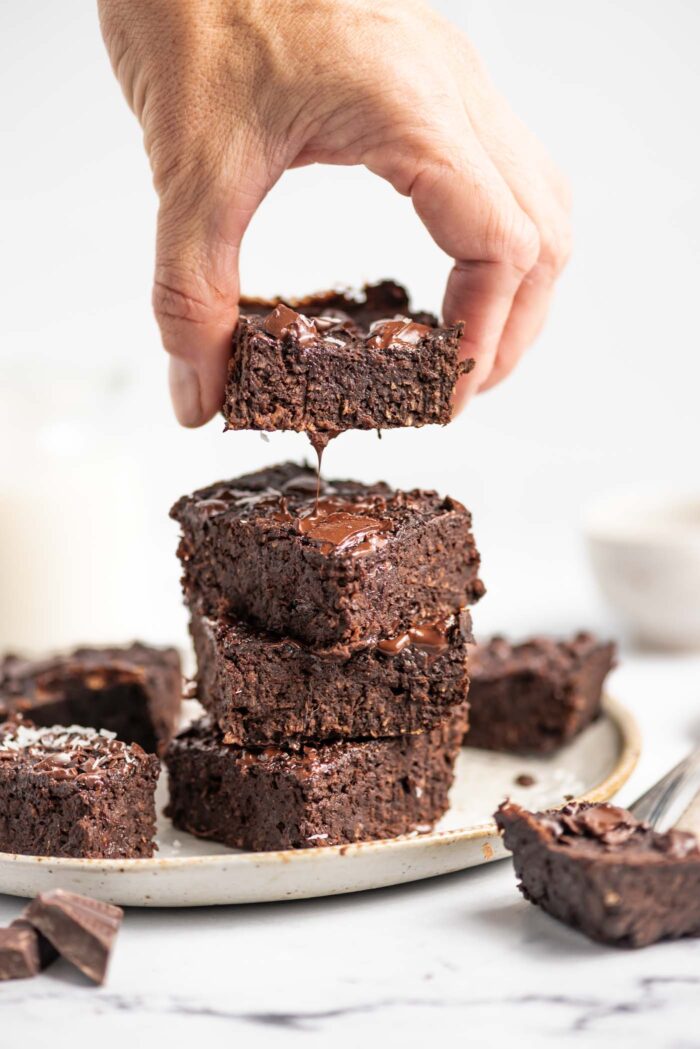 Hand lifting a chocolate brownie from a stack of 4 brownies on a plate.