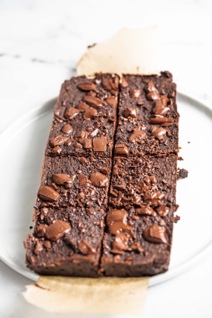 A slab of coconut brownies cut into 8 portions topped with chocolate chunks on a piece of parchment paper on a plate.
