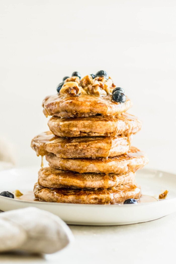 Stack of fluffy vegan pancakes topped with blueberries, walnuts and maple syrup on a plate.