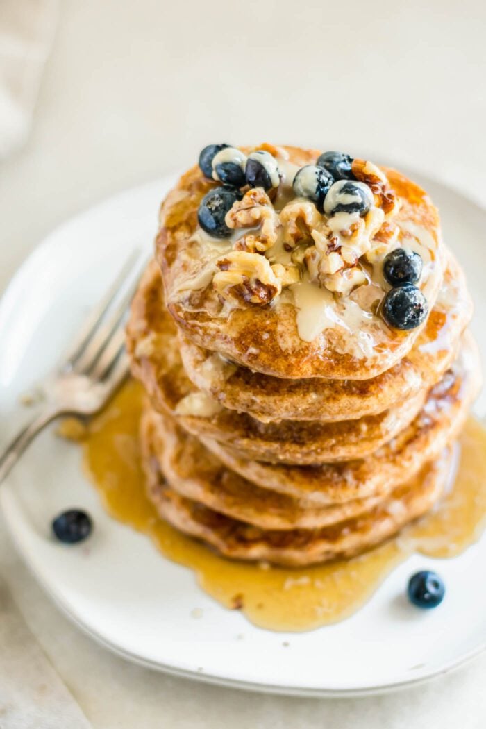 Stack of fluffy whole wheat pancakes topped with walnuts, tahini, maple syrup and blueberries on a plate with a fork.