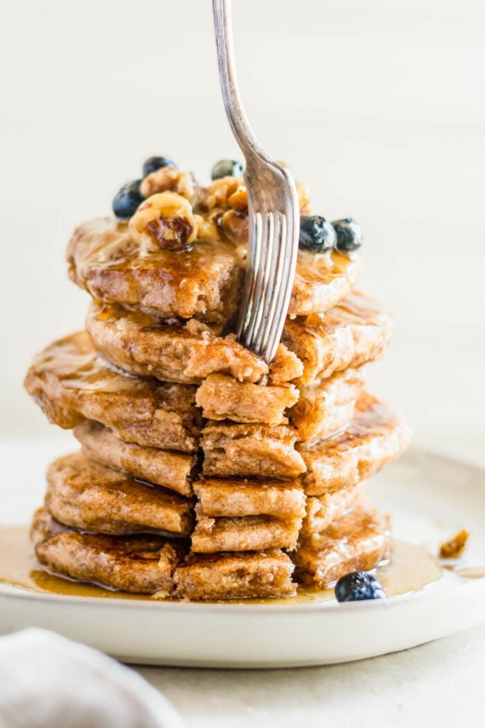 Fork digging into a big stack of whole wheat pancakes on a plate. The pancakes are topped with walnuts, blueberries and maple syrup.