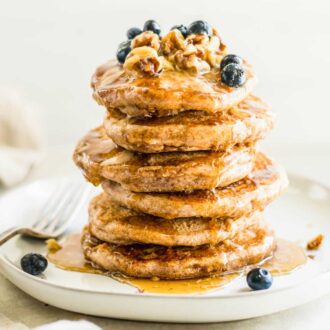 Stack of fluffy vegan pancakes topped with blueberries, walnuts and maple syrup on a plate. A fork rests on the plate.