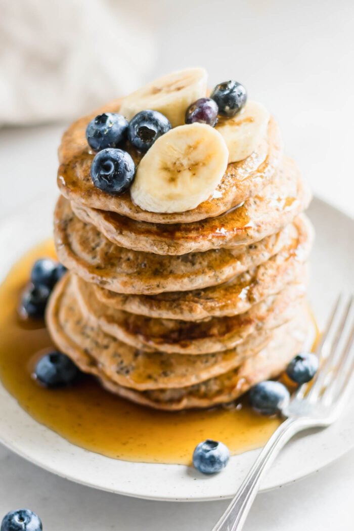 Stack of chia pancakes topped with blueberries, banana and maple syrup on a plate with a fork.