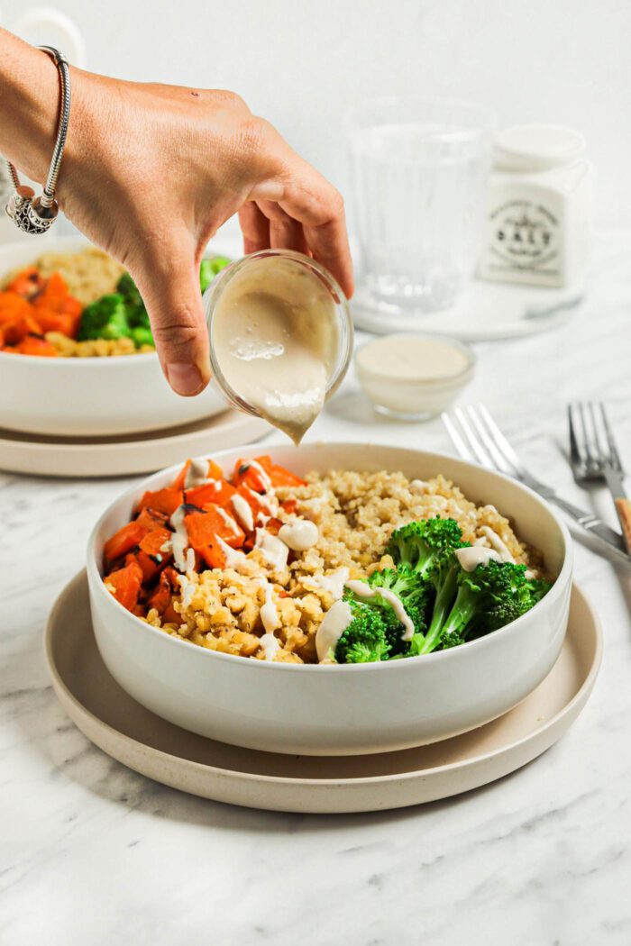 Hand pouring tahini sauce from a small jar over a bowl with lentils, quinoa, roasted vegetables and broccoli.