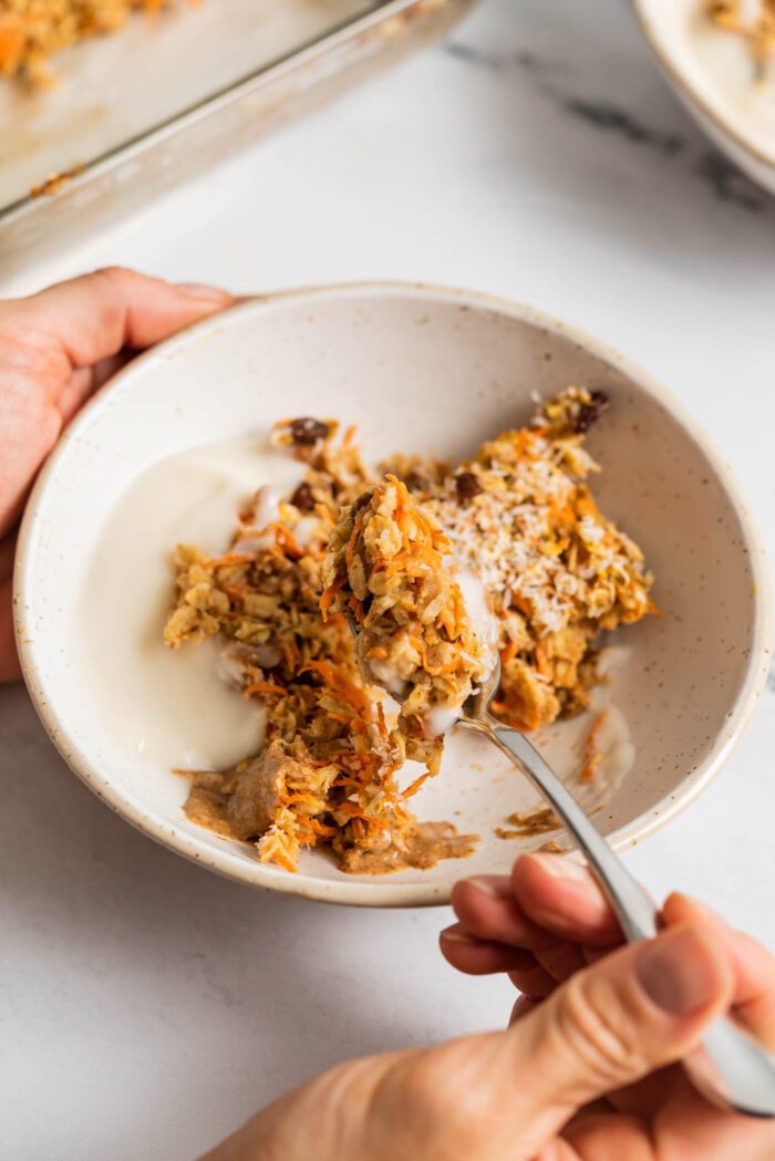 Hand using a spoon to lift a spoonful of carrot cake baked oatmeal from a bowl with yogurt and almond butter.