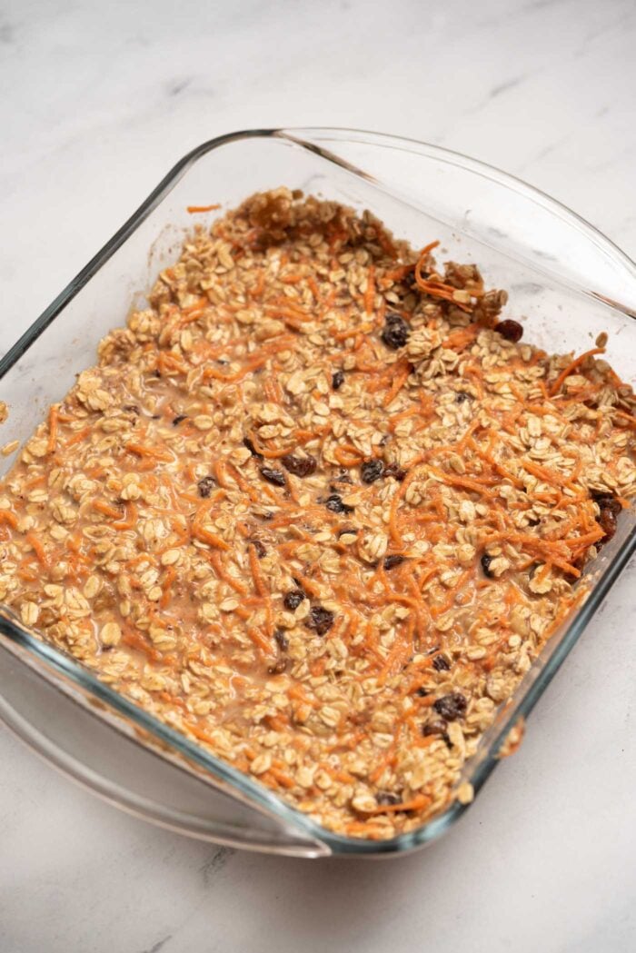 Rolled oats mixed with walnuts, raisins and grated carrot in a square baking dish.