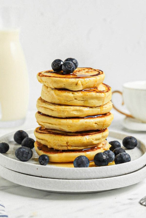 Stack of 6 thick chickpea flour pancakes topped with blueberries and maple syrup on a small plate. A jug of milk and a coffee cup are in the background.