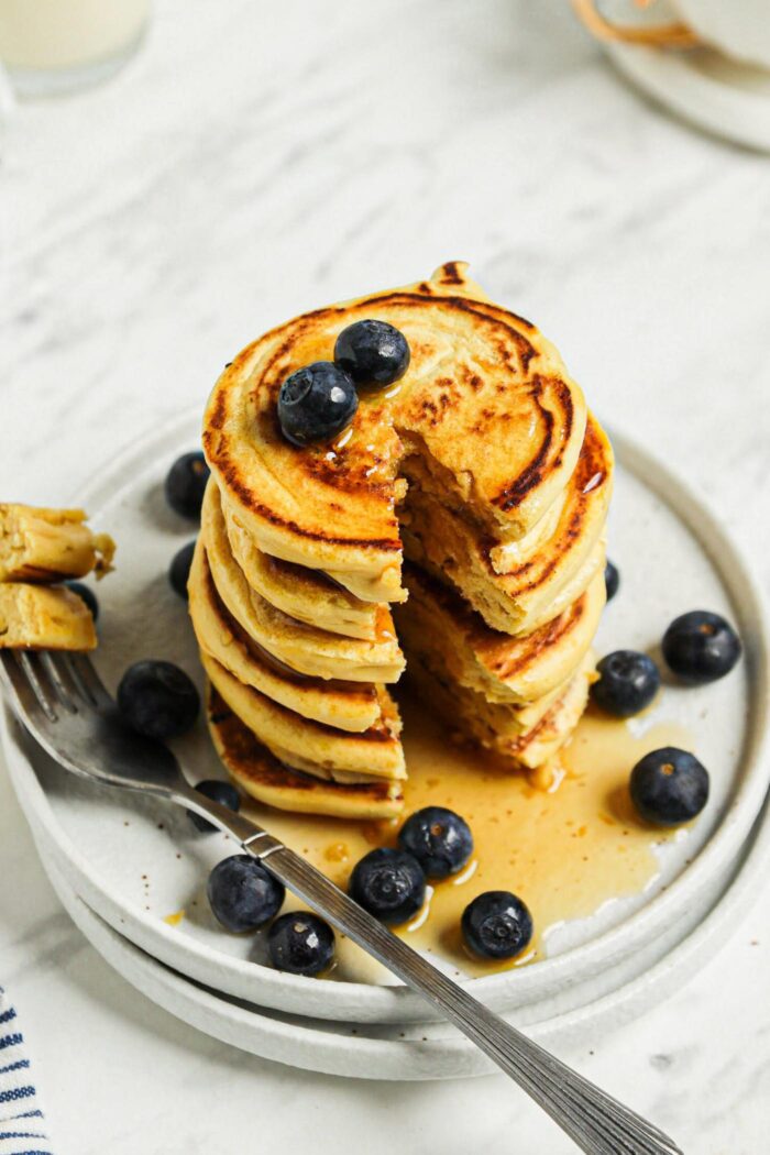 A large stack of chickpea blueberry pancakes on a plate with a slice taken from them by a fork.