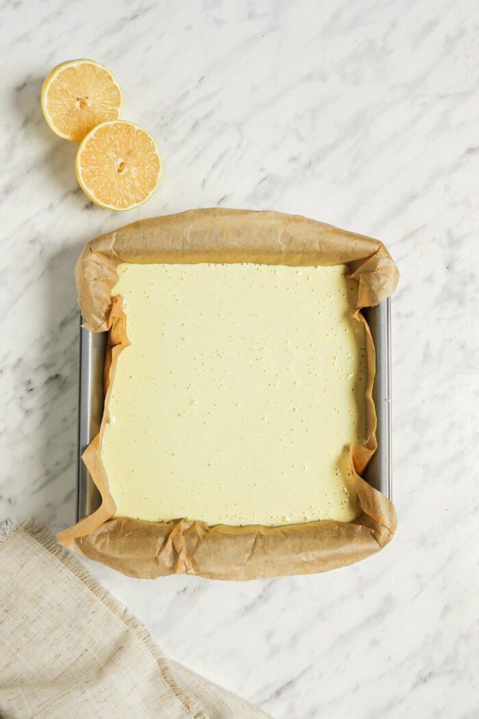 Overhead view of an unbaked lemon bar in a square baking pan lined with parchment paper. 2 slices of lemon are beside the pan.