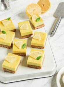 Numerous vegan lemon squares on an upside down baking tray on a marble surface. Slice of lemon and a lemon zester are in the background.