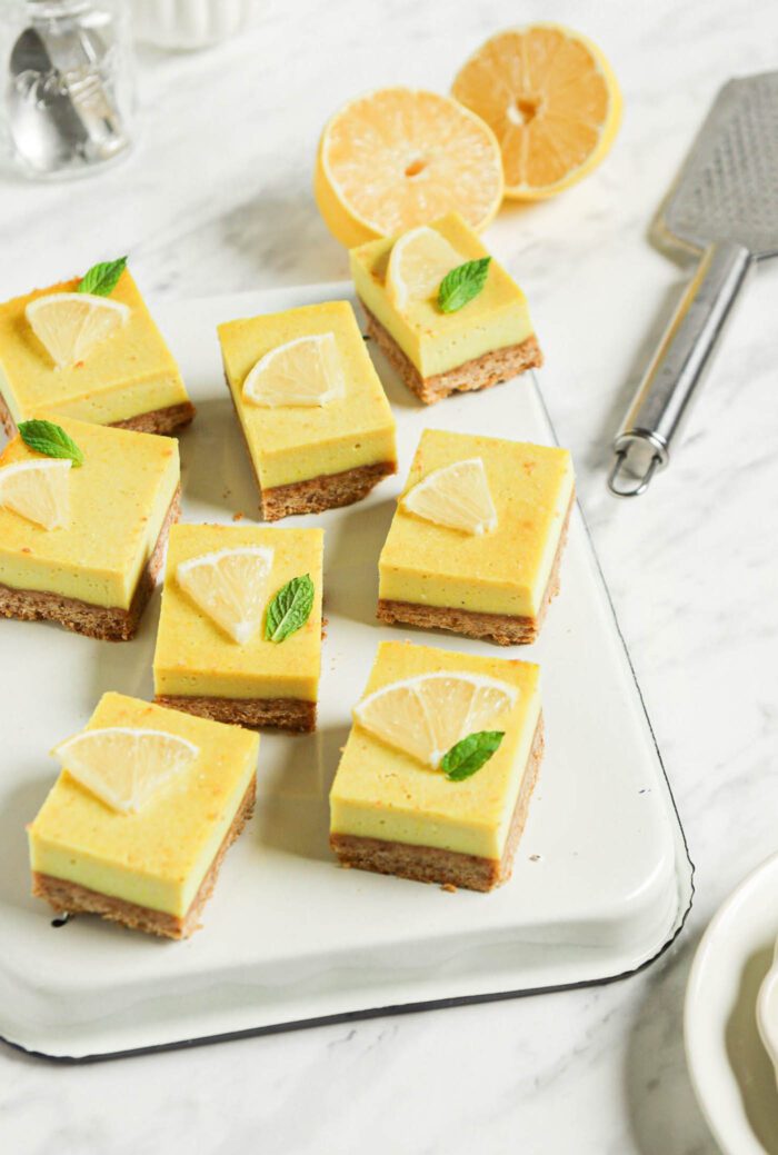 Numerous vegan lemon squares on an upside down baking tray on a marble surface. Slice of lemon and a lemon zester are in the background.