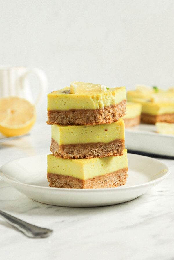 Stack of 3 creamy vegan lemon bars on a small white plate on a marble countertop.