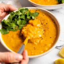 Hand using a spoon to lift a spoonful of lemon lentil soup topped with fresh cilantro from a bowl. Another bow of soup is in the background.