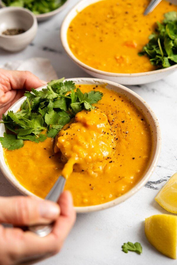 Hand using a spoon to lift a spoonful of lemon lentil soup topped with fresh cilantro from a bowl. Another bow of soup is in the background.