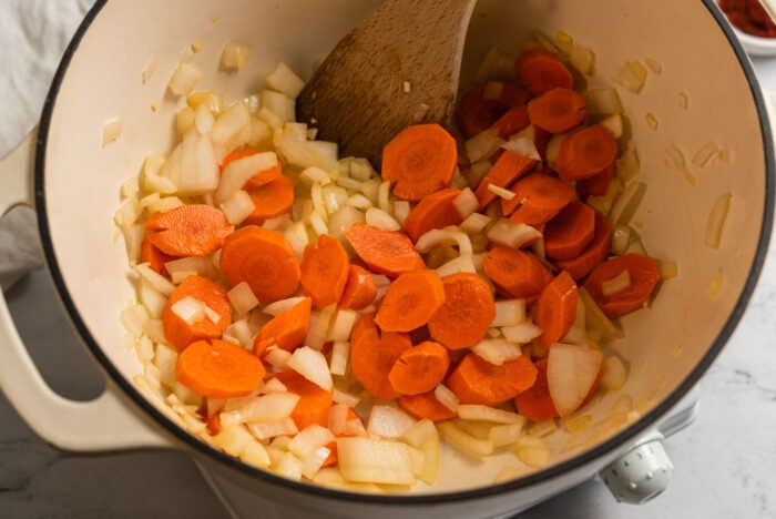 Onion, garlic and carrot cooking in a large soup pot with a wooden spoon.