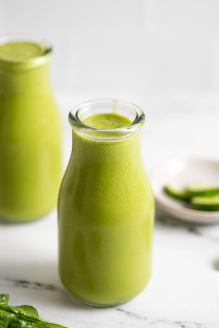 Two jars of creamy green mango pineapple smoothie with a few pieces of spinach scattered around them.