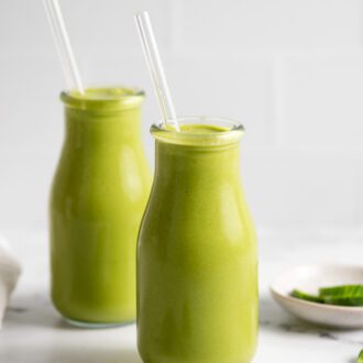 Two jars of green smoothie with glass straws in them. A few pieces of spinach are scattered around the jars.