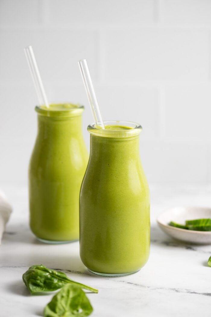 Two jars of green smoothie with glass straws in them. A few pieces of spinach are scattered around the jars.