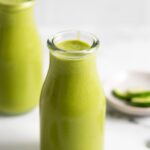 Two jars of creamy green mango pineapple smoothie with a few pieces of spinach scattered around them.