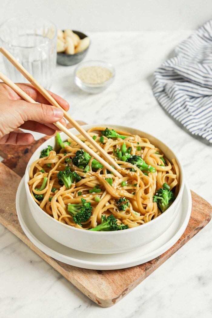 Bowl of sesame noodles with broccoli topped with scallions and sesame seeds. A pair of chopsticks rests on the bowl.