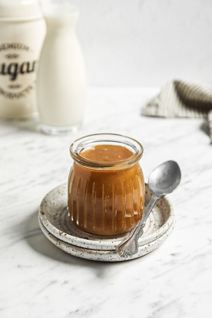 Small jar of homemade caramel sauce on a plate with a spoon beside it.