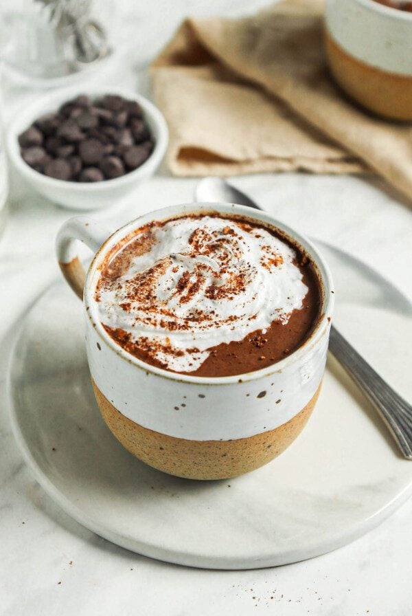 Close up of a mug of hot chocolate topped with whipped cream on a small plate with a spoon and a small whisk.