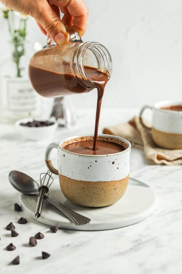 Pouring hot chocolate from a glass jar into a mug.