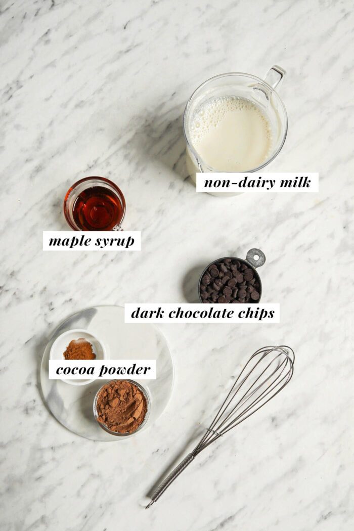 Maple syrup, milk, chocolate chips and cocoa powder in bowls on a marble counter.