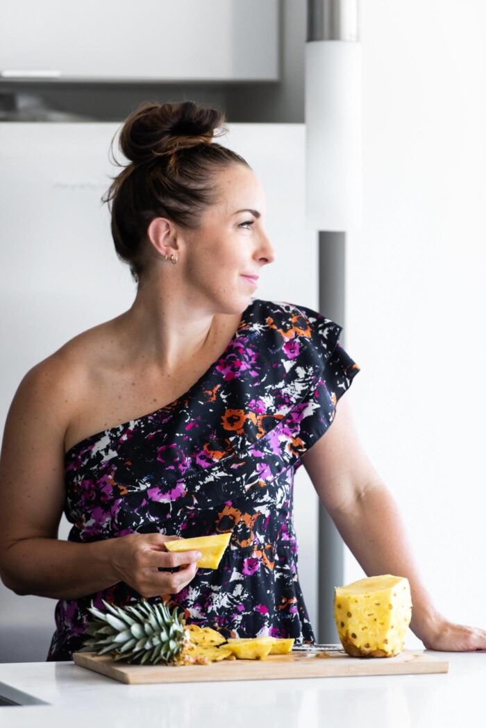 A woman wearing a one-shoulder floral dress looking over her shoulder holding a slice of fresh pineapple over a cutting board with more sliced pineapple on it.