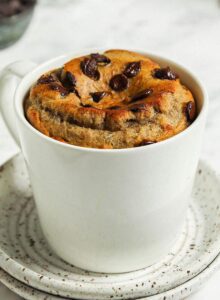 Microwave-baked banana bread mug cake with chocolate chips in a mug sitting on two small plates.