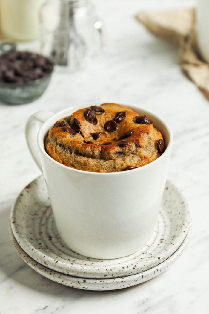 Microwave-baked banana bread mug cake with chocolate chips in a mug sitting on two small plates.