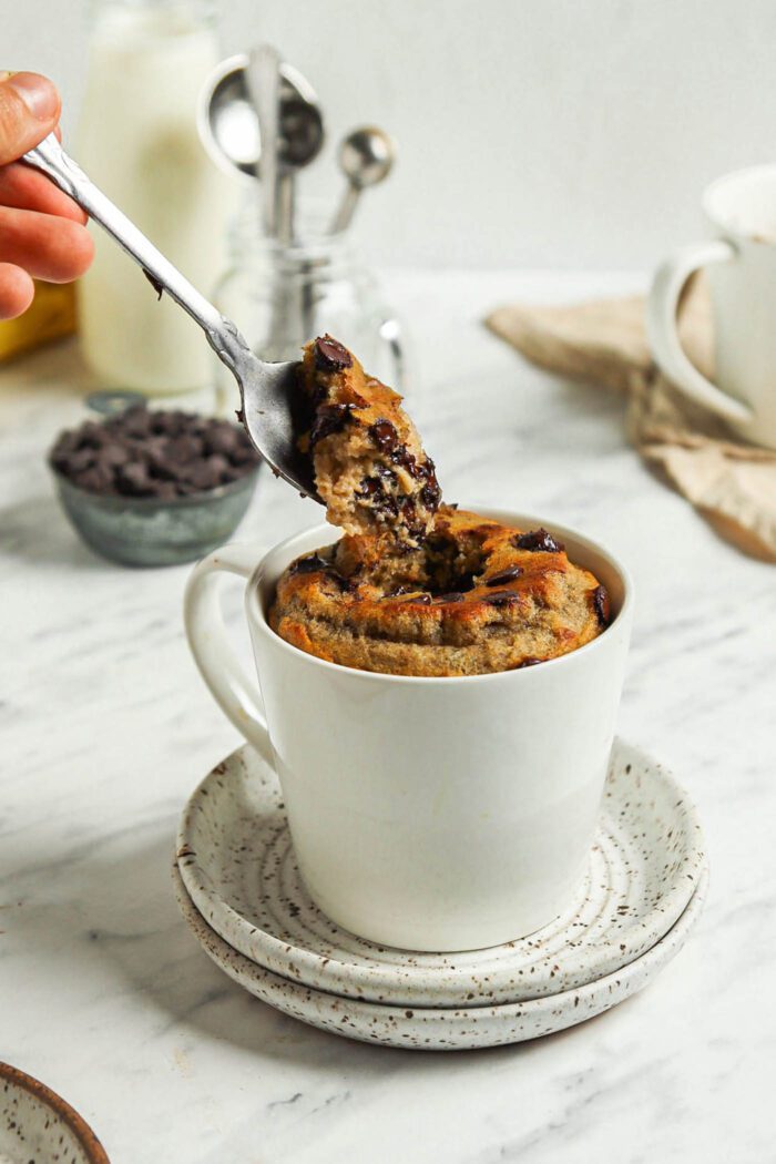 Spoon lifting a spoonful of banana bread mug cake from a mug sitting on two small plates.