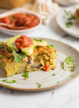 Slice of vegan breakfast casserole with potato and veggies topped with avocado and salsa. Hand is using a fork to take a slice from it.