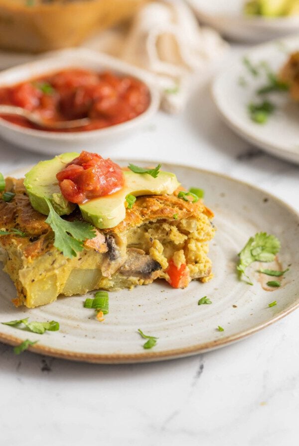 Slice of vegan breakfast casserole with potato and veggies topped with avocado and salsa. Hand is using a fork to take a slice from it.