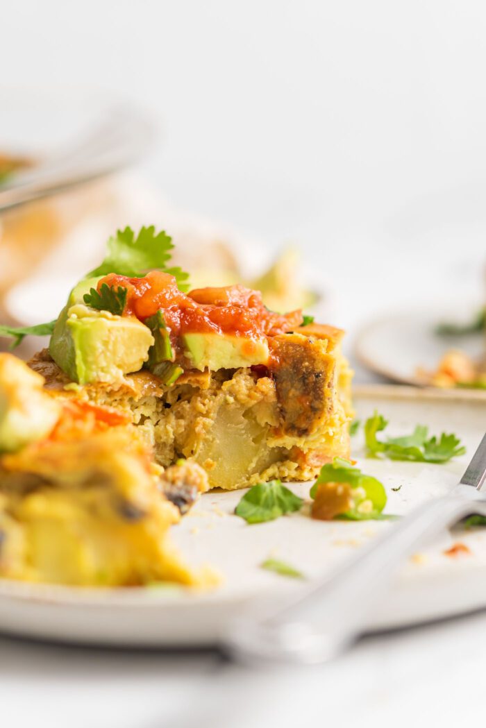 Close up of a slice of vegan eggless casserole with chunks of potato and veggies in it, topped with avocado and salsa.