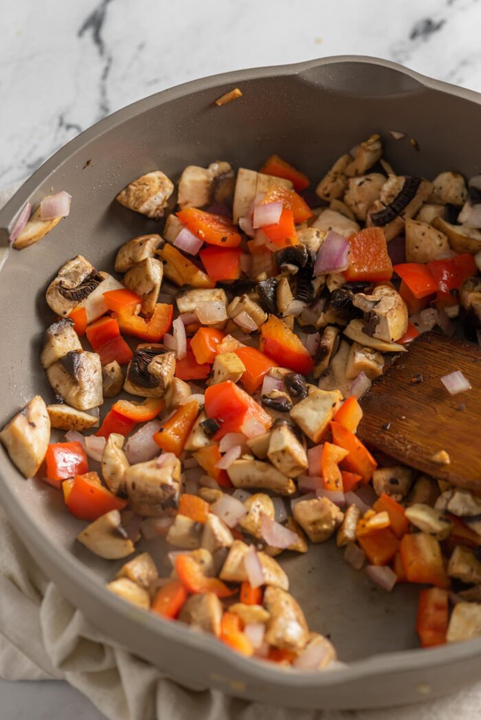 Mushroom, onion and red bell pepper cooking in a skillet with a wooden spoon.
