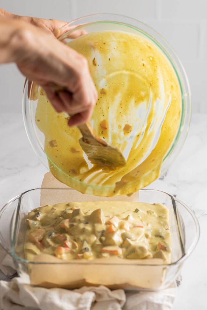 Pouring egg potato casserole batter from a bowl into a square glass baking pan.