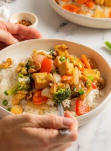Hands using a fork for cashew tofu stir fry over rice and topped with green onions served on plate.