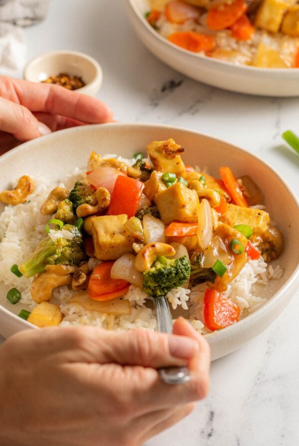 Hands using a fork for cashew tofu stir fry over rice and topped with green onions served on plate.