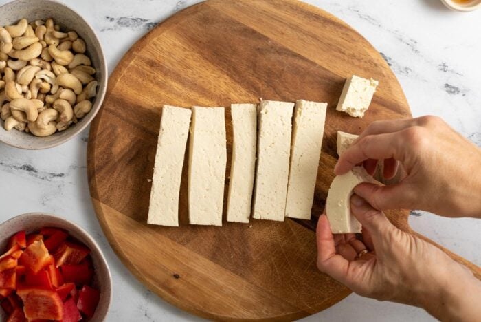 Strips of tofu on a cutting board. Two hands breaking the strips into small cubes.