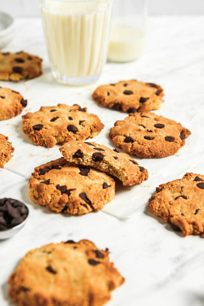 Numerous almond flour chocolate chip cookies on a marble surface with a glass of milk in the background.