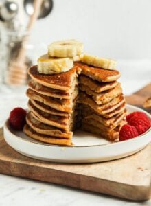Stack of vegan banana oatmeal pancakes with a slice taken out of them so you can see the texture inside. The pancakes are topped with sliced banana and raspberries.