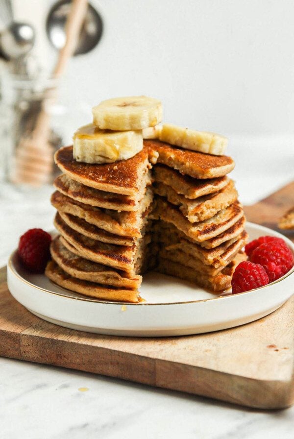 Stack of vegan banana oatmeal pancakes with a slice taken out of them so you can see the texture inside. The pancakes are topped with sliced banana and raspberries.