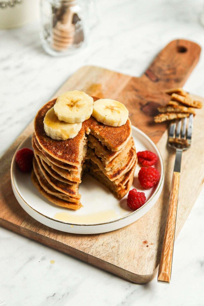 Overhead view of a stack of vegan banana oatmeal pancakes with a slice taken out of them so you can see the texture inside. The pancakes are topped with sliced banana and raspberries and on a plate sitting on a cutting board.