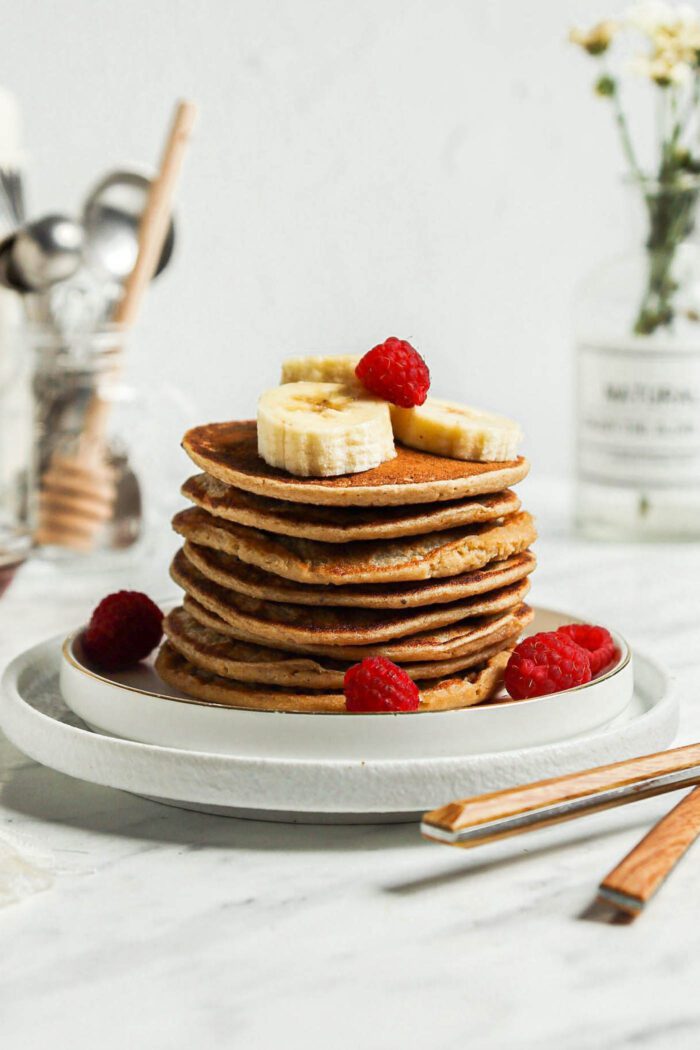 Stack of healthy vegan banana oatmeal pancakes on a plate topped with sliced bananas and fresh raspberries.