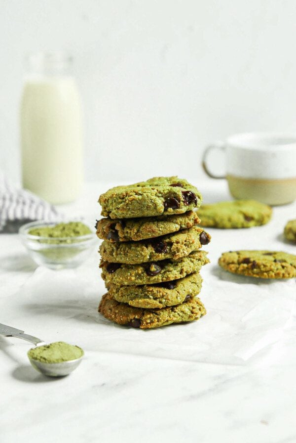 Stack of almond flour matcha chocolate chip cookies. There's a glass of milk in the background and a tablespoon of matcha powder in the foreground.