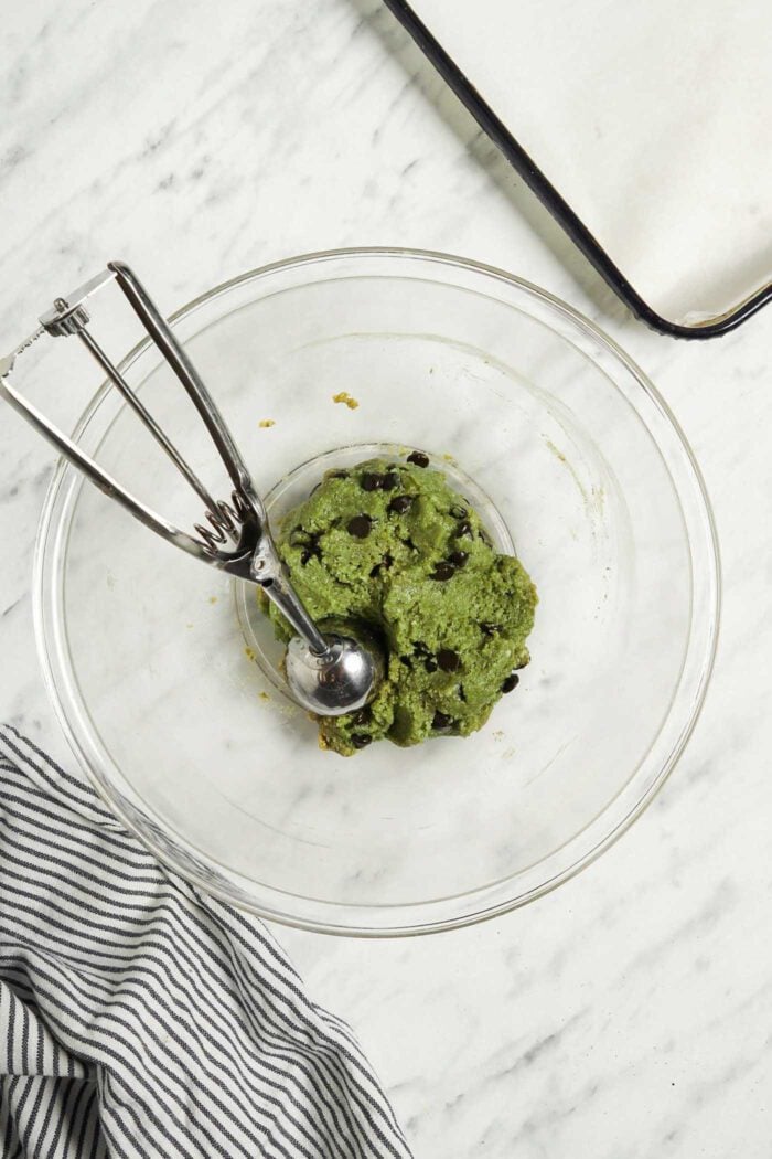 Raw vegan matcha cookie dough in a glass mixing bowl with a cookie scoop..