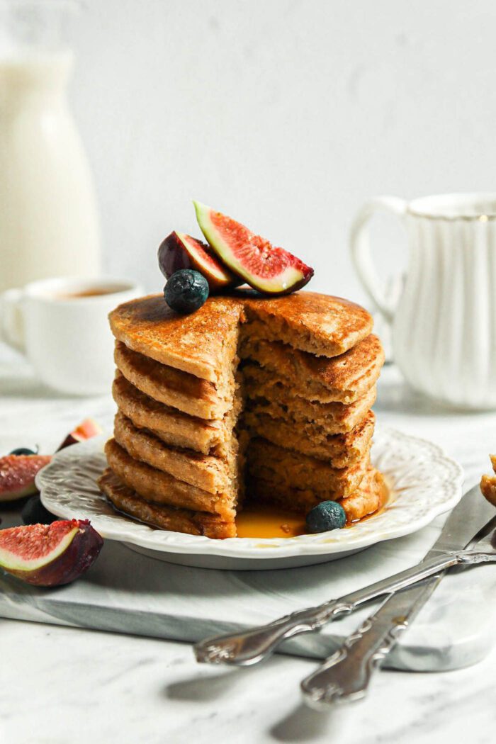 Stack of thick vegan oat flour pancakes topped with blueberries and figs on a small plate.The stack has a slice taken from it so you can see the inside texture of the pancakes.