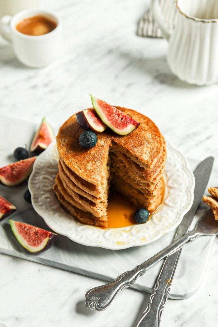 Stack of thick vegan oat flour pancakes topped with maple syrup, blueberries and figs on a small plate.The stack has a slice taken from it so you can see the inside texture of the pancakes.