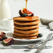 Hand pouring maple syrup from a small container over a stack of healthy oatmeal pancakes.